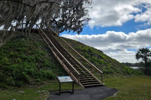 Manatees And More: 25 Things To Do In Crystal River, Florida 2025 21 Wooden staircase ascending a historic native american burial mound covered with greenery, under a cloudy blue sky. An information plaque is visible at the mound's base, inviting visitors to learn about this sacred site.