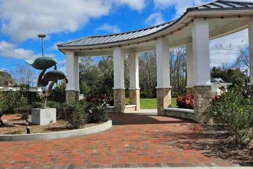 Is Crystal River Worth Visiting In 2025 11 White pergola with classical columns stands in a park, framing a whimsical metal sculpture of manatees in the foreground. The brick pathway leading to the structure invites visitors to enjoy a moment of leisure under the clear blue sky, surrounded by well-maintained shrubbery