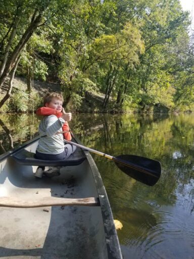 Best Campgrounds Near Philadelphia, Pa For 2025 7 A child in a life jacket gives a thumbs up while sitting in the bow of a canoe, with an oar in the water. The canoe is surrounded by lush green trees reflecting off the calm water.