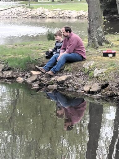Best Campgrounds Near Philadelphia, Pa For 2025 5 An adult and a child are sitting by a pond, fishing together. Their peaceful reflection can be seen on the water's surface, along with the greenery and rocks that line the pond.