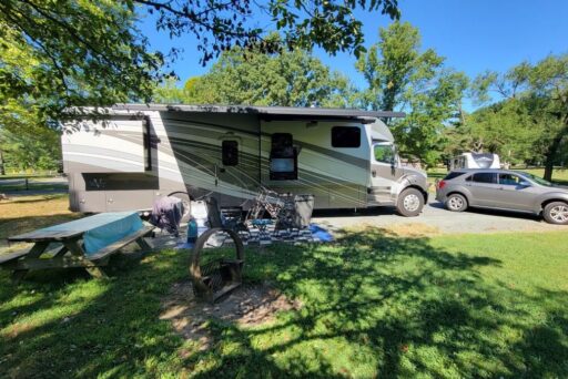 Best Campgrounds Near Philadelphia, Pa For 2025 3 A large rv parked at a sunny campsite with a picnic table, fire pit, and bicycles. Nearby, a car is hitched to a portable satellite dish under a canopy of trees with dappled sunlight filtering through.