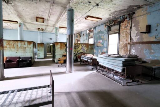 Best Campgrounds Near Philadelphia, Pa For 2025 21 Interior of an abandoned institution with peeling paint on the walls and ceiling, featuring rows of old metal-framed beds and a solitary bench in the foreground, evoking a sense of desolation.