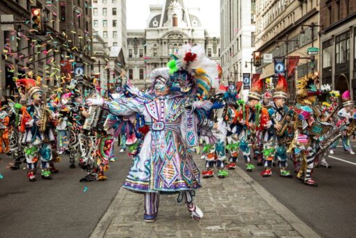 Best Campgrounds Near Philadelphia, Pa For 2025 18 A vibrant mummers parade in progress, with participants clad in elaborate, colorful costumes and feathered headdresses, playing instruments and dancing on a city street scattered with confetti, with historic architecture in the background