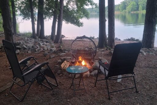 Easy Rv Trip Planning: Preparing For Your Next Adventure 11 Two camping chairs face a small campfire by a lakeside, surrounded by trees and rocky ground. A small table between the chairs holds two glasses of wine, creating a cozy and serene outdoor setting. The calm lake and lush greenery in the background add to the peaceful atmosphere.