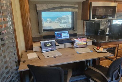 Rv Apps: Which Ones Do You Need? 12 A workspace inside an rv. This setup includes multiple laptops on a spacious desk, positioned by a window, providing a bright and comfortable working environment. The modern kitchen area in the background adds to the convenience of working on the road. This image perfectly captures the essence of balancing work and travel, making it clear that an rv can serve as a fully functional mobile office. Whether you're a digital nomad or just need to stay connected while traveling, this setup shows how easily you can work remotely from your rv.