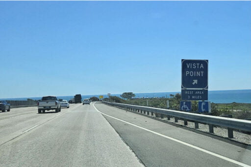 Rv Apps: Which Ones Do You Need? 3 The image shows a highway running alongside the ocean under a clear blue sky. Several vehicles are driving along the road. On the right side of the road, there is a large blue sign that reads,