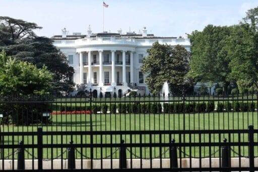 Rv Friendly Campgrounds Near Washington, Dc 2025 16 The image shows the white house in washington, d. C. , viewed from behind a black iron fence. The iconic building stands with its distinctive white columns and curved portico, surrounded by lush green lawns, manicured bushes, and mature trees. A small fountain sprays water in front of the white house, with the american flag flying atop the roof.