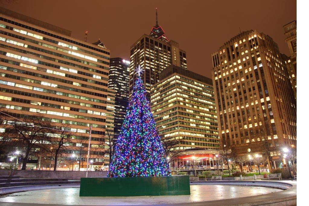 A beautifully lit Christmas tree stands tall in a Philadelphia plaza, surrounded by towering buildings with bright windows against the night sky. The tree sparkles with multicolored lights, adding festive cheer to the urban landscape. Perfect for celebrating Christmas in Philadelphia.