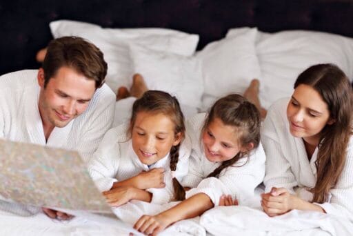Swim With Manatees In 2025: An Unforgettable Adventure 9 A family of four wearing white robes, lying on a bed and smiling while looking at a map. The parents and two daughters appear engaged and happy, suggesting a moment of planning or bonding during a relaxing getaway.