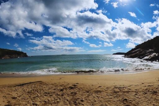 Beaches In Acadia National Park: More Than Just Sand 4 Sandy beach with gentle waves, rocky cliffs, and a bright blue sky with scattered clouds.