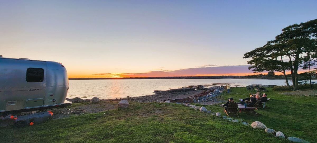 This image showcases a peaceful waterfront campsite at sunset, with a silver Airstream trailer parked on the left. The golden sun dips below the horizon, casting a warm glow over the calm waters, while a group of people relaxes in Adirondack chairs near a fire pit on the grassy shore.