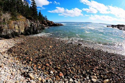 Beaches In Acadia National Park: More Than Just Sand 8 Little hunters beach with a rocky shoreline, ocean waves, and a forested cliff under a bright blue sky.