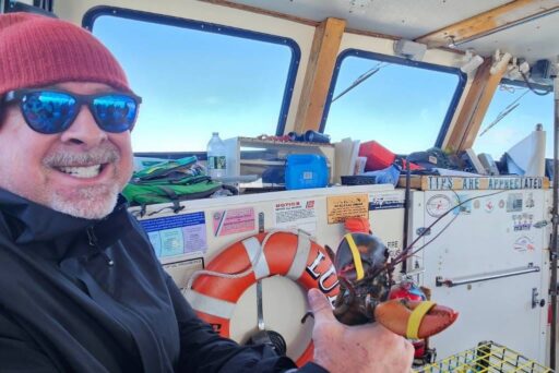The Ultimate Acadia National Park Itinerary For 2025: Your Perfect 3-Day Plan 20 A smiling man in sunglasses and a beanie holds a live lobster aboard a lobster boat, surrounded by fishing gear and ocean views.