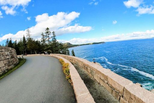 The Ultimate Acadia National Park Itinerary For 2025: Your Perfect 3-Day Plan 4 A scenic stretch of park loop road in acadia, curving along a stone wall with ocean views, forested hills, and a bright blue sky.