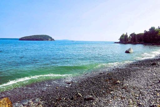 Beaches In Acadia National Park: More Than Just Sand 25 A rocky beach in bar harbor with gentle waves, a boulder in the water, and an island in the distance.