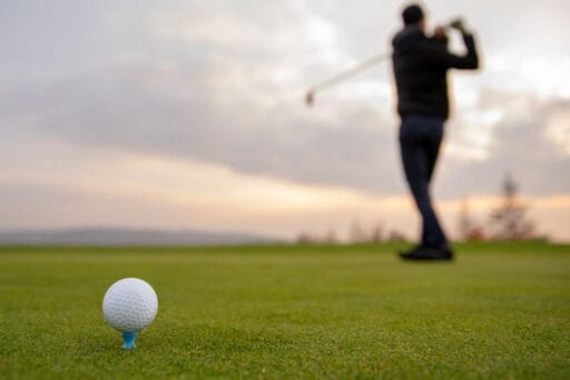30 Best Things To Do In Batavia, Ny In 2025 10 Close-up of a golf ball on a tee with a golfer swinging in the background at a course in batavia, ny, during sunset.