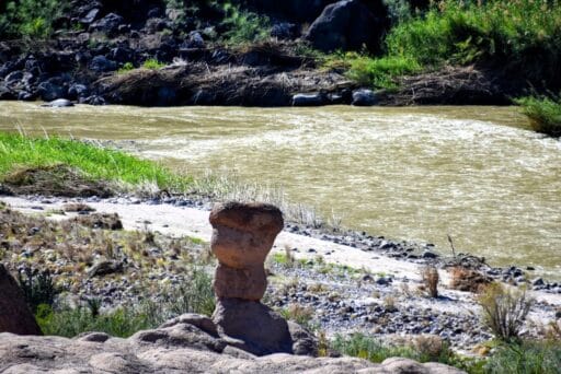 11 Fun Things To Do In Terlingua You Can’t Miss In 2025 13 Stacked rock hoodoo in the foreground beside a muddy river, with green vegetation and rocky terrain along the banks.