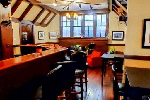 30 Best Things To Do In Batavia, Ny In 2025 12 Cozy speakeasy-style tavern room with dark wood beams, bar seating, framed art, and red lounge chairs near large paned windows.