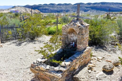 11 Fun Things To Do In Terlingua You Can’t Miss In 2025 3 Old stone grave with wooden cross in terlingua cemetery, surrounded by desert plants and distant mountains.