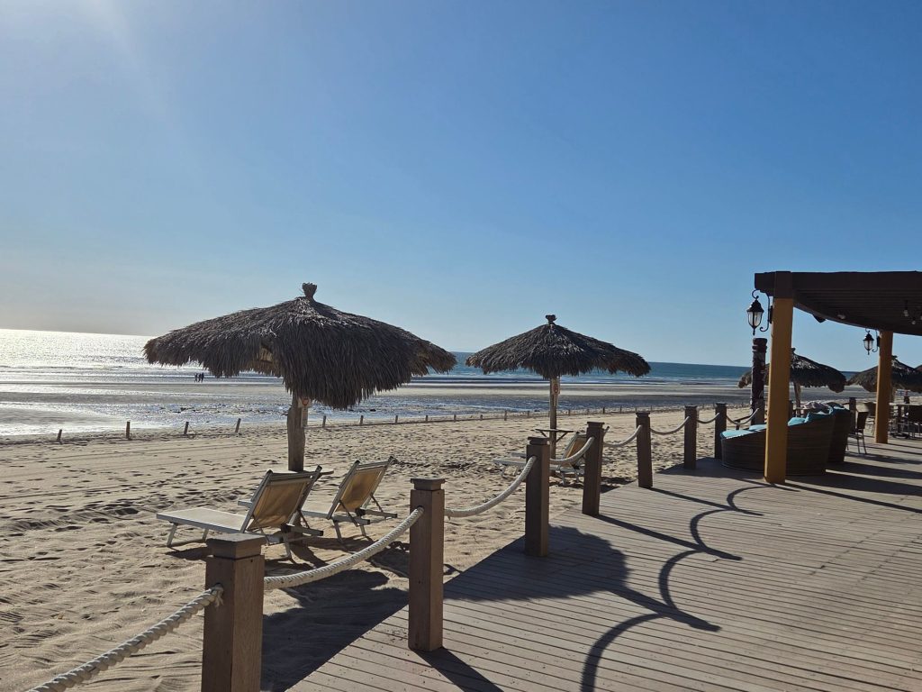 Beach loungers under thatched umbrellas facing the ocean in puerto peñasco with soft morning light reflecting off the water. The quiet and relaxed setting captures the kind of slow beach days that make it easy to stay longer in puerto peñasco.