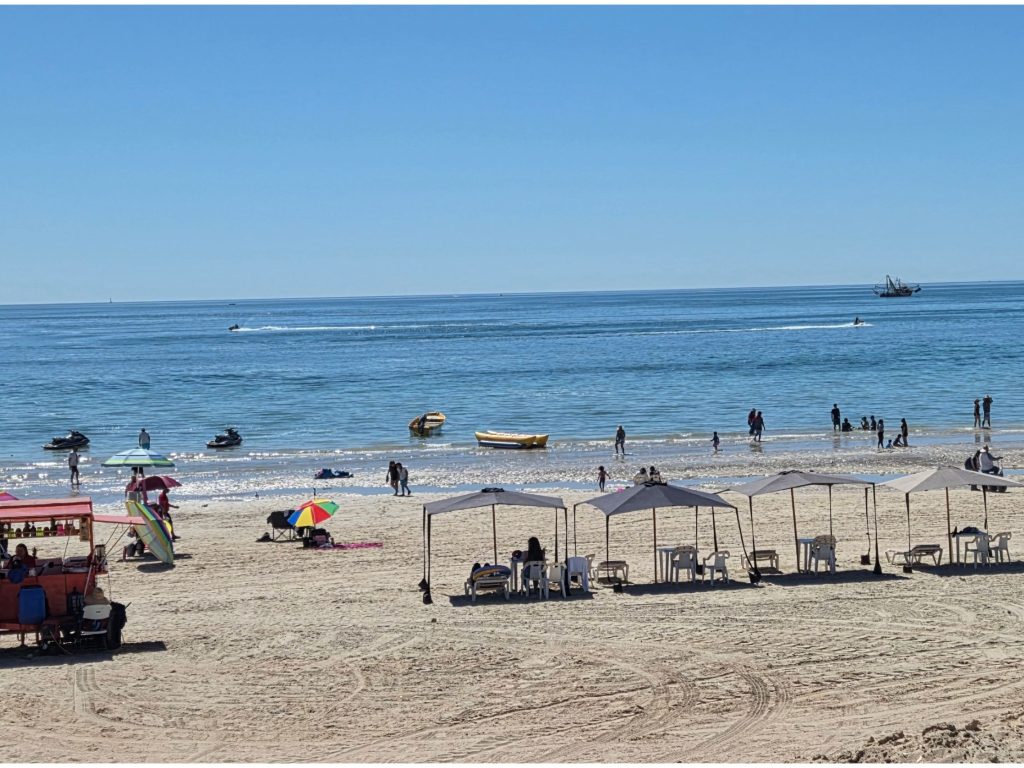People relaxing under shaded umbrellas on sandy beach in puerto peñasco while others swim and ride jet skis along the shoreline. This lively beach scene highlights why relaxing on sandy beach is one of the best things to do in puerto peñasco on a sunny day.
