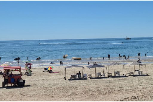People relaxing under shaded umbrellas on sandy beach in puerto peñasco while others swim and ride jet skis along the shoreline. This lively beach scene highlights why relaxing on sandy beach is one of the best things to do in puerto peñasco on a sunny day.