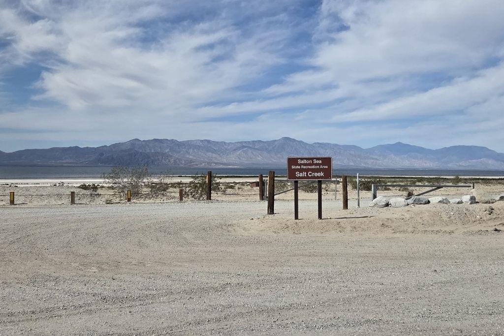 Visiting The Salton Sea: California’s Strangest Desert Lake 2 Salt creek entrance sign beside the salton sea with desert mountains and dramatic clouds behind it.