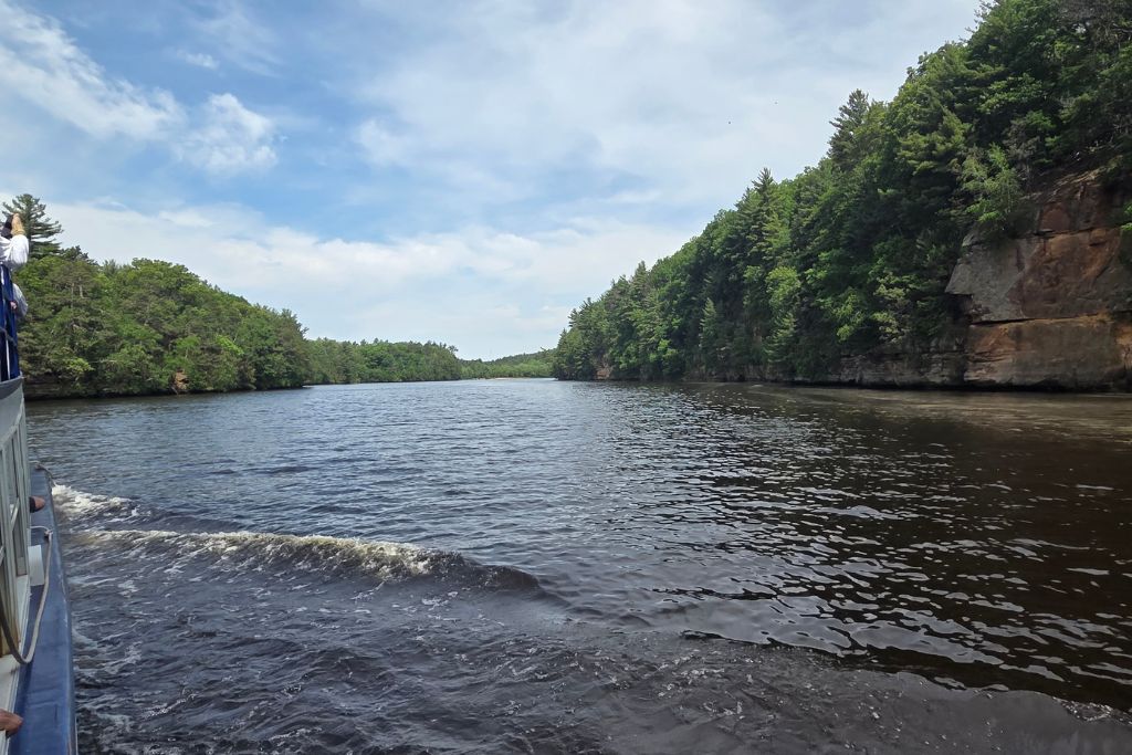 Wisconsin Dells For Adults: Beyond The Waterparks 3 View from an upper dells boat tour cruising along the wisconsin river with forested sandstone cliffs rising beside the dark water. Passengers can be seen along the side of the tour boat as it travels through the scenic upper dells.
