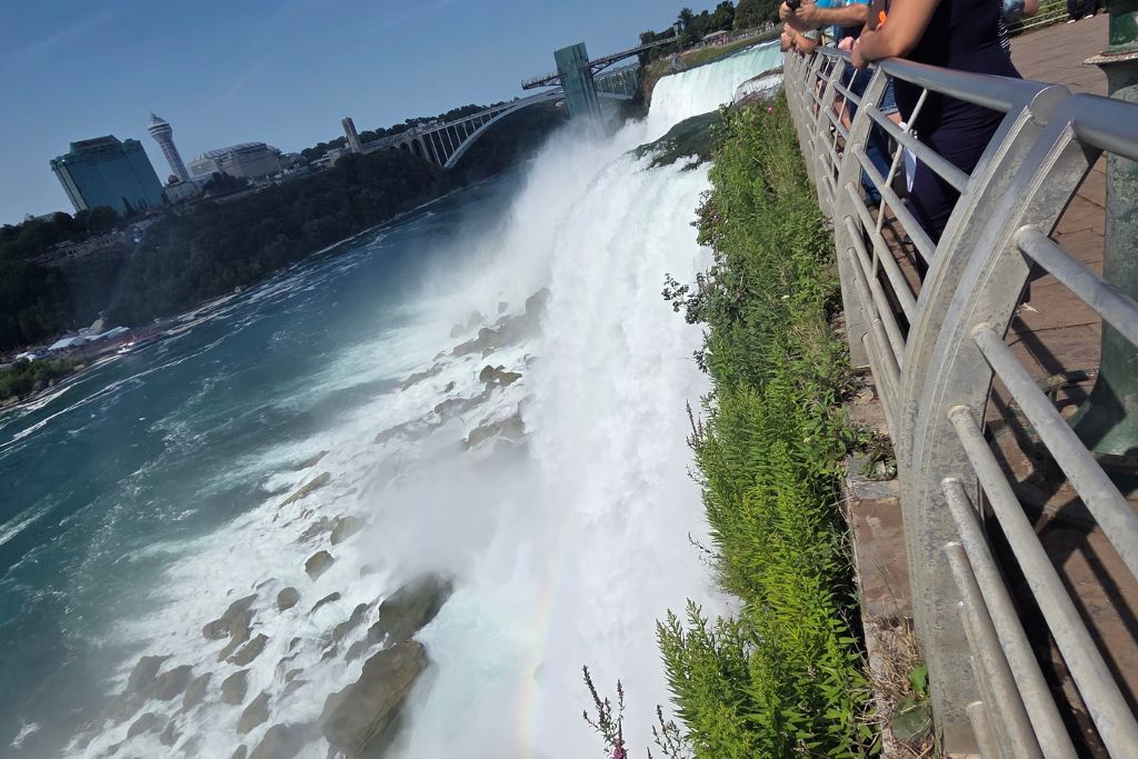 Is Niagara Falls Worth Visiting? What We Think After Several Visits 5 American falls plunges beside the railing at the luna island viewing platform as a maid of the mist boat travels through the niagara river below. Visitors stand only a few feet from the rushing water with mist and a faint rainbow rising from the rocks at the base of the falls.