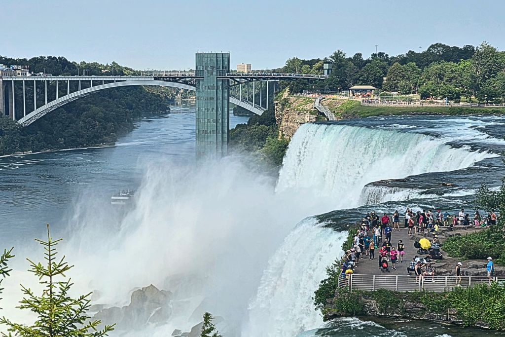 Luna Island From Stedmans Bluff At Niagara Falls American falls and bridal veil falls plunge beside luna island as visitors crowd the viewing platform just feet from the rushing water. Rainbow bridge spans the niagara river in the background in this view from stedmans bluff on the american side.