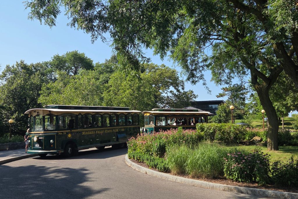 Is Niagara Falls Worth Visiting? What We Think After Several Visits 3 Two green niagara falls state park trolleys travel along a tree shaded road surrounded by gardens on a sunny summer day. The trolley makes it easier to travel between niagara falls viewpoints and attractions without walking the entire park.