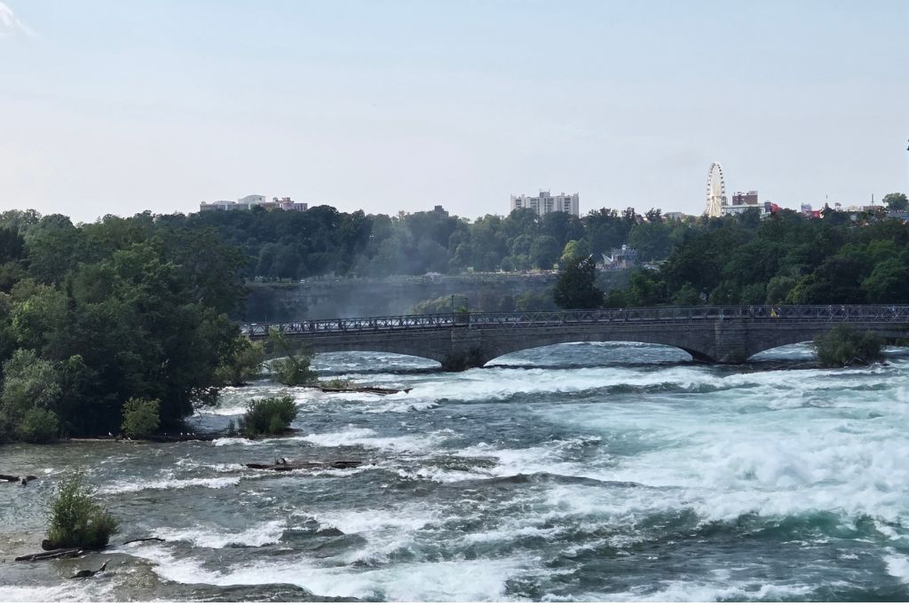 Is Niagara Falls Worth Visiting? What We Think After Several Visits 7 Whitewater rapids churn across the niagara river as the water races past small tree covered islands and beneath a stone pedestrian bridge. The turbulent water shows the force of the niagara river as it approaches the falls.