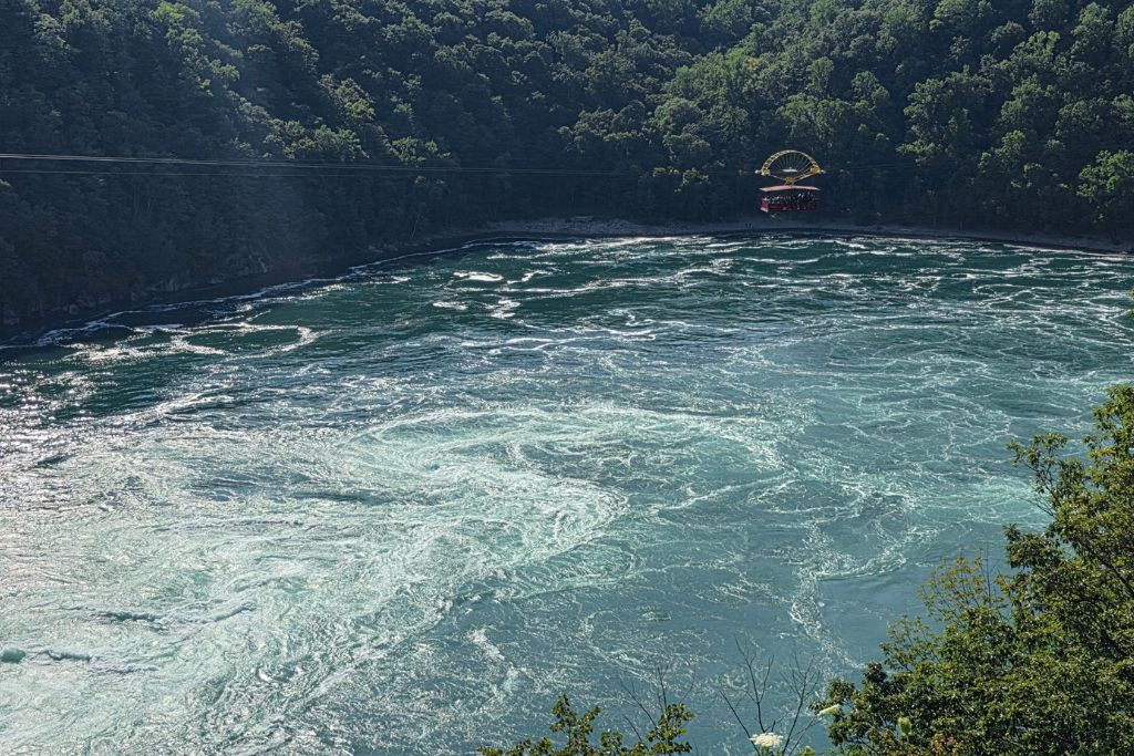 Is Niagara Falls Worth Visiting? What We Think After Several Visits 8 Aqua blue water swirls through the niagara whirlpool below the steep forested walls of the niagara gorge. The red whirlpool aero car hangs above the churning water in the distance while crossing from the canadian side.