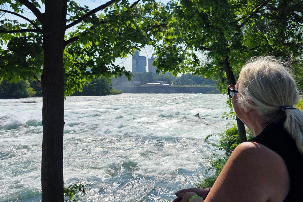 Is Niagara Falls Worth Visiting? What We Think After Several Visits 14 Maureen stands beneath leafy shade trees watching the powerful niagara river rapids rush past in niagara falls state park. The quiet moment beside the water captures a different side of visiting niagara falls after a day spent exploring the overlooks.