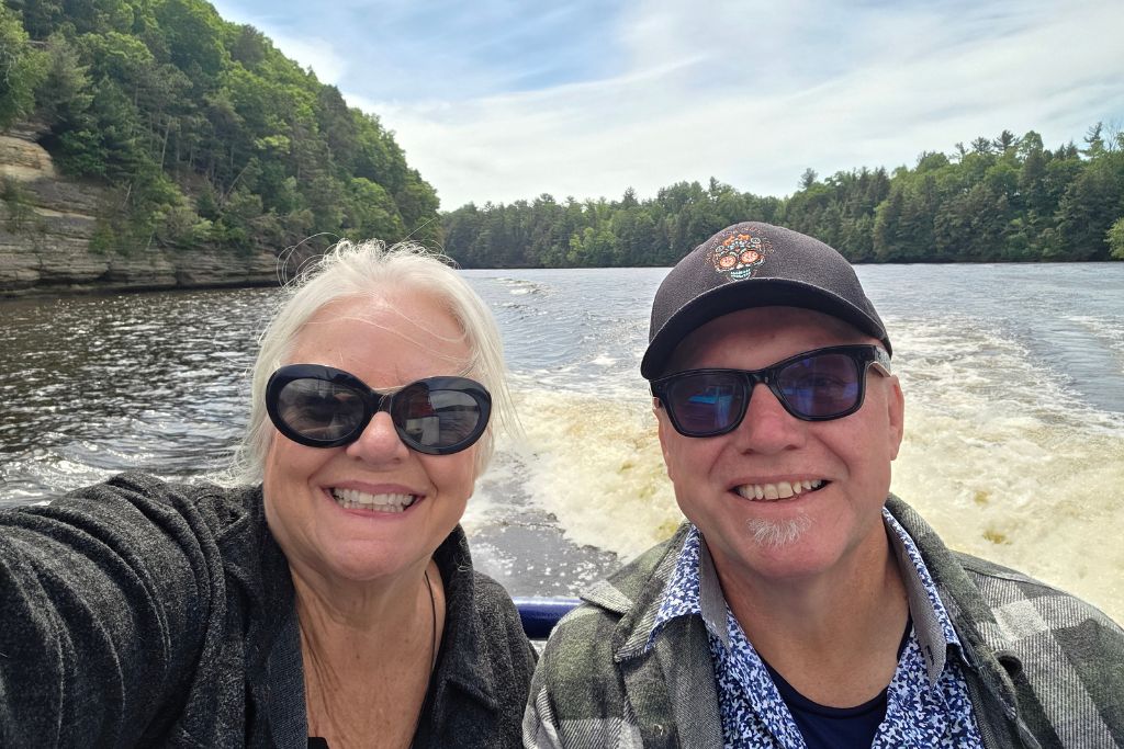 Wisconsin Dells For Adults: Beyond The Waterparks 19 A smiling couple takes a selfie during a boat tour through the forested sandstone cliffs of the wisconsin dells. The river and wake from the boat stretch behind them.